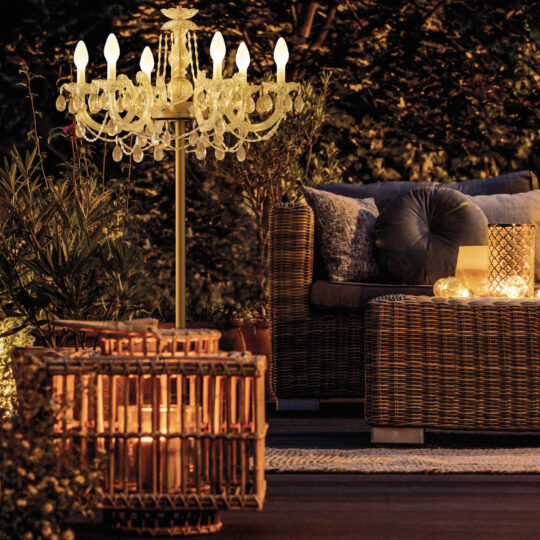 A cozy night-time outdoor seating area features wicker furniture with cushions. An Outdoor Crystal Chandelier Table Lamp hangs above, casting a gentle glow, while decorative candleholders add warmth. Surrounding plants create a serene ambiance.
