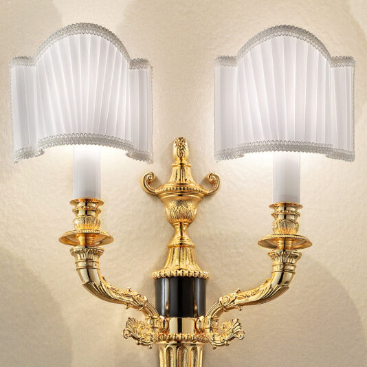 The Classic Gold Plated Double Wall Light is an ornate fixture with two elegantly curved arms, each adorned with a white pleated lampshade. Intricate detailing and a central decorative finial enhance its classic appeal, creating a soft glow against a beige, textured background.