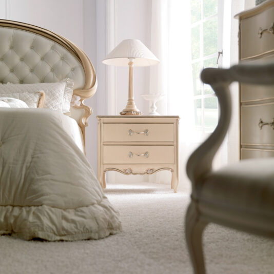 Classic Italian Rose Design Vermeil Bedside Table An elegant bedroom features a large bed with a tufted, curved headboard, plush bedding, and a soft, neutral color palette. The Classic Italian Rose Design Vermeil Bedside Table, adorned with a lamp and decorative items, stands beside the bed while natural light streams through a nearby window.