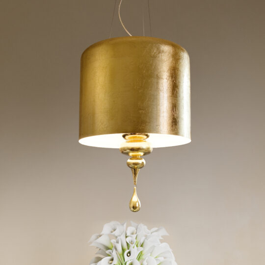 A Modern Italian Designer Gold Leaf Ceiling Light with a cylindrical shade and ornate drop detail illuminates white calla lilies against a warm background.