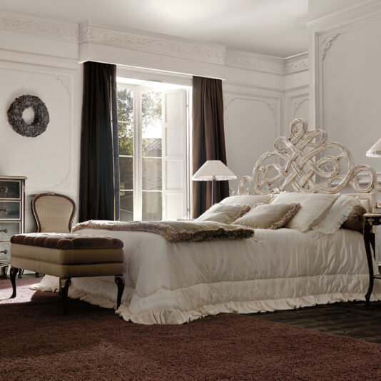 Designer Italian Button Upholstered Storage Bench Elegant bedroom with a large ornate bed, a Designer Italian Button Upholstered Storage Bench at the foot, and two bedside lamps. Tall windows with dark curtains let in natural light. The room features white walls, crown molding, a rug, a chest of drawers, and a decorative wreath.