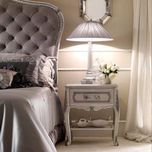 A luxurious bedroom showcases a tufted headboard, the Ornate Italian Bedside Table adorned with intricate carvings, a table lamp featuring a pleated shade, a vase of white flowers, and a decorative bowl on the lower shelf. The decor is dominated by soft, neutral tones, creating an ambiance of serene elegance.