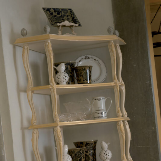 An Ornate Italian Designer Corner Shelf, in a decorative cream-colored wooden finish with four levels, displays various items including ceramic vases, decorative plates, cups, and a small teapot against a white wall. This piece features intricate carved details along its edges.