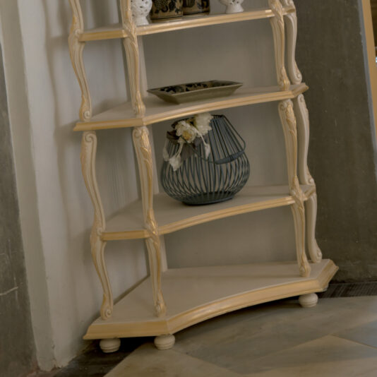 Against a wall stands an Ornate Italian Designer Corner Shelf, featuring curved legs and four tiers. The second tier is adorned with a decorative wire vase filled with white flowers, while the third tier supports a small dish. Beneath the shelf, the floor showcases a pattern of light and dark diagonal tiles.