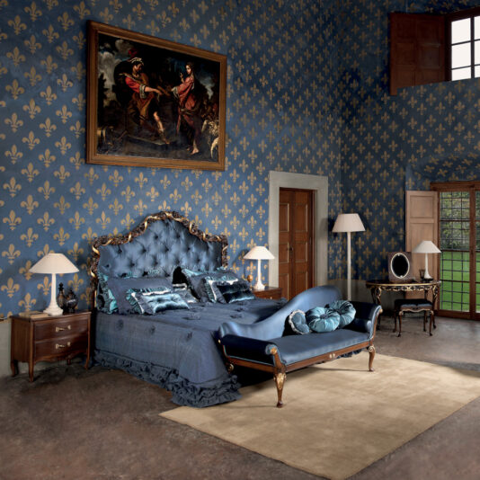 Elegant bedroom featuring a luxurious tufted bed with blue bedding and matching pillows. The room has blue wallpaper with gold fleur-de-lis patterns, complemented by classic wooden furniture. A Reproduction Two Tone Walnut Italian Chaise Longue adds a touch of timeless luxury, while tall windows enhance the sophisticated decor.