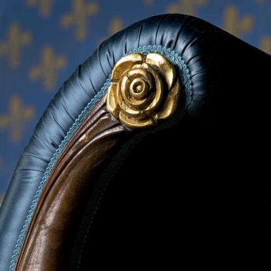 Close-up of an ornate chair detail featuring a gold rose accent at the top corner. The black fabric is trimmed with a plush turquoise edging, and the dark wooden frame of this Reproduction Two Tone Walnut Italian Chaise Longue is partially visible. The background shows a blue pattern with gold fleur-de-lis motifs.