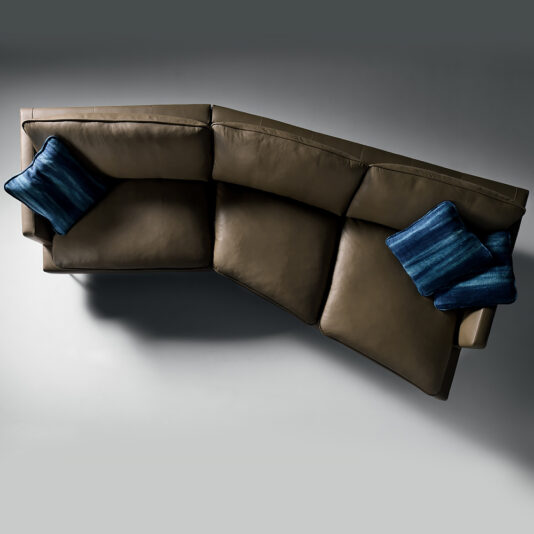 Top view of the Modern Leather Modular Sofa, featuring an L-shaped design with three seat cushions and two blue decorative pillows. The brown leather sectional sits against a plain gray background, casting a shadow on the floor.