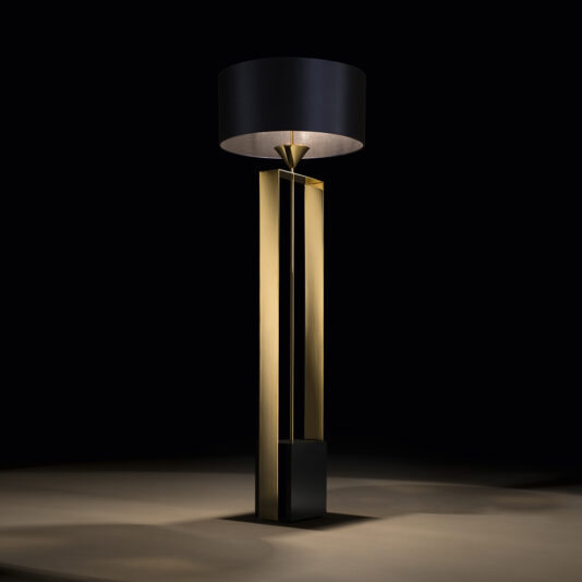The Gold Contemporary Designer Floor Lamp features a sophisticated, modern design with a tall, rectangular gold base and an oversized, cylindrical black lampshade that emits a soft, warm light. This exquisite piece stands against a dark background, casting a subtle glow on the floor.