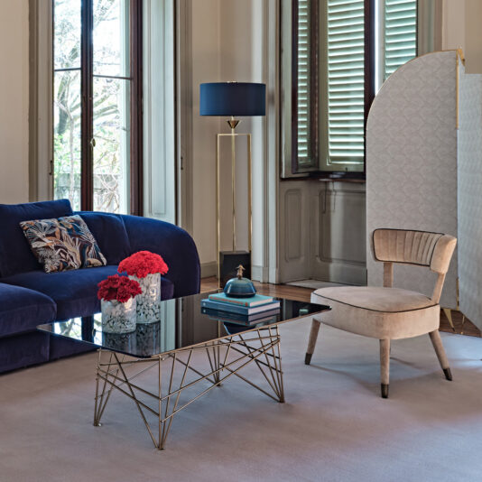 A modern living room featuring a blue velvet sofa, a beige accent chair, and a glass-top coffee table with a geometric metal base. The table holds vibrant red flowers and books. In the background are tall windows with shutters, the Gold Contemporary Designer Floor Lamp, and a partition screen.