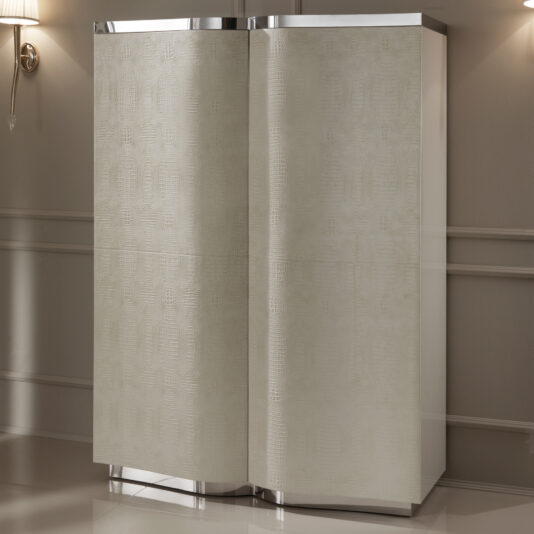The Textured Leather Italian Designer Large Cabinet, in modern beige with a subtle leather pattern and chrome accents on the top and bottom, stands against a neutral wall; a wall-mounted lamp is partially visible to the left.
