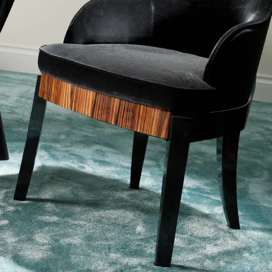Designer Italian Macassar Velvet Dining Chair The Designer Italian Macassar Velvet Dining Chair showcases curved armrests, four angled legs, and a wooden Macassar accent on the front edge in sleek black, all elegantly positioned on a soft teal carpet.