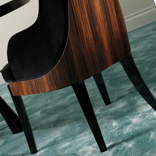 Designer Italian Macassar Velvet Dining Chair The Designer Italian Macassar Velvet Dining Chair, featuring a curved wooden backrest and elegant black upholstery, rests on a light teal carpet. Its black legs complement the luxurious design, while a corner of a refined black table is visible beside it.