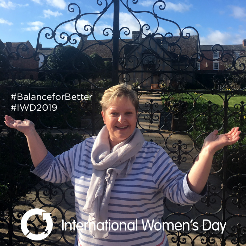 A person stands outdoors in front of a decorative iron gate, smiling with arms raised. They are wearing a striped shirt and scarf. Overlay text reads "#BalanceforBetter #IWD2019" and "International Women’s Day" with its logo. The sky is blue with scattered clouds.