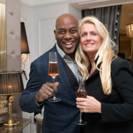 A smiling man and woman holding glasses of sparkling wine, posing together in a warmly lit room with elegant decor including lamps and chandeliers. The man is wearing a dark blue checkered suit, and the woman is in a black outfit with long blonde hair.