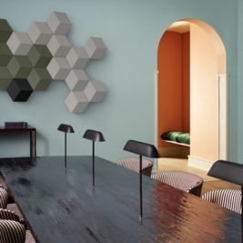 A modern conference room with a long wooden table, surrounded by striped chairs. The wall is adorned with hexagonal 3D art pieces. A bookshelf stands on the left, and an archway on the right leads to a nook with a green cushion. The walls are painted blue and peach.