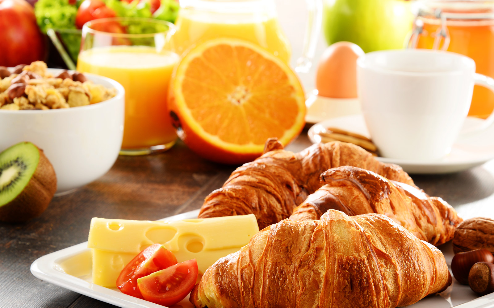 Be the perfect host with a breakfast spread featuring two croissants, cheese slices, cherry tomato halves, and a plate of kiwi. Accompanied by a bowl of cereal with nuts, a glass of orange juice, a white coffee cup, a whole orange, and various fruits in the background.