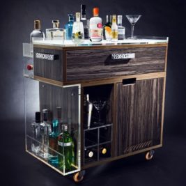 A sleek, modern bar cart with various bottles of alcohol neatly arranged on top and in shelves. It has a wooden finish with metallic accents and is equipped with wheels for mobility. Perfect for World Gin Day, the cart includes a drawer, a glass section, and space for bar tools and glasses.