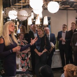 A woman in a dark blue dress speaks to an attentive audience in an elegantly decorated room with modern lighting. The audience, dressed in formal attire, listens intently. The atmosphere suggests a formal event or gathering.