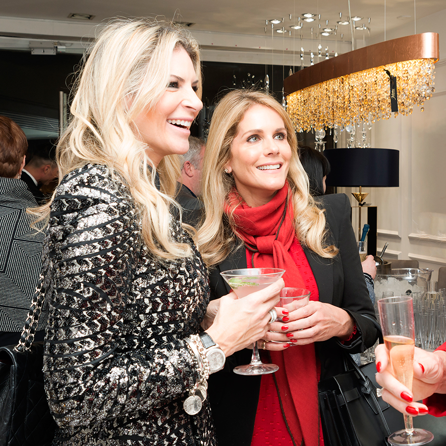 Two women are joyfully conversing at a social event. One wears a shimmering, metallic outfit and holds a martini while the other, in a black blazer and red scarf, holds a glass of champagne. Both have long blonde hair and are smiling. The setting is elegant and festive.