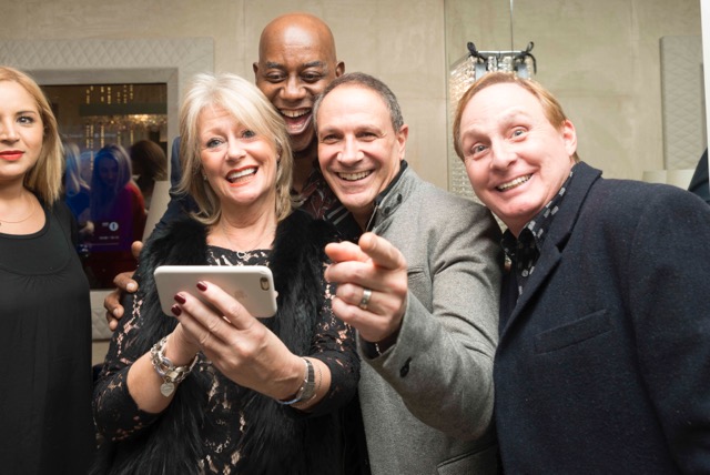 A group of four people are joyfully gathered together. One woman in front is holding a smartphone to take a selfie. The others, including another woman and two men, are smiling and laughing towards the camera. They appear to be at a social event or gathering.