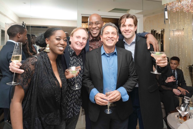 A group of five people dressed in formal attire stand closely together at what appears to be a party or social event. They are smiling and holding drinks. The background features elegant decor, including a chandelier and a large mirror, and other attendees are visible.