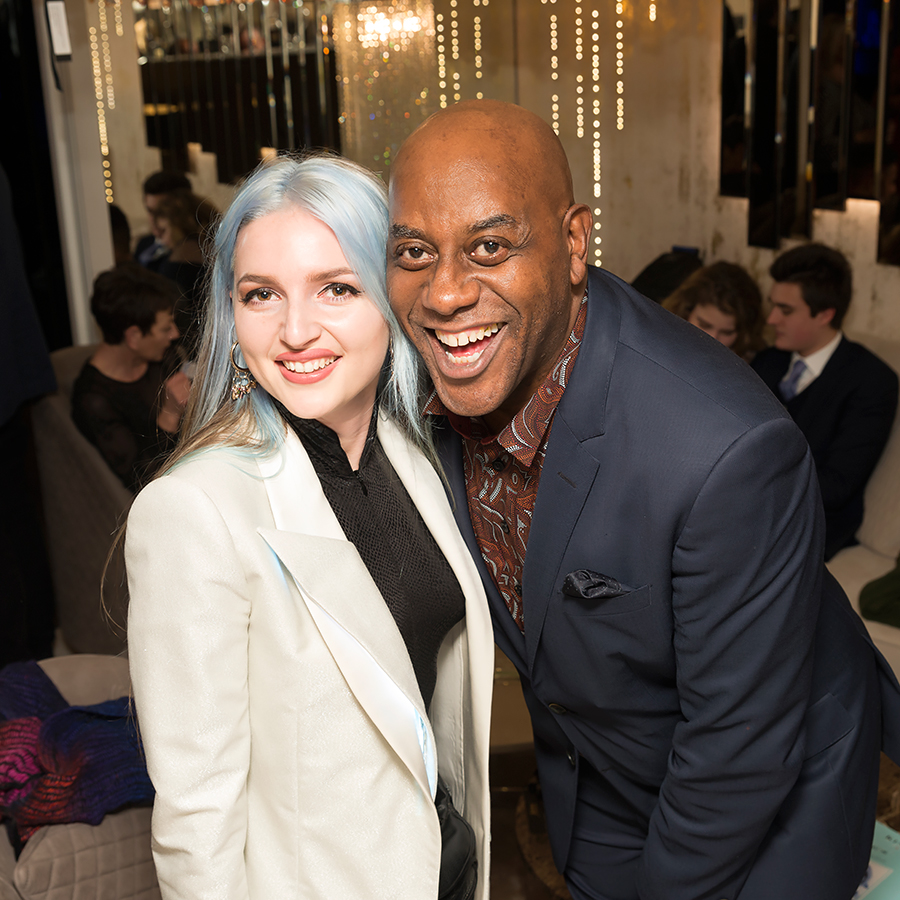 Two people, dressed formally, are smiling at the camera in an indoor setting with other people in the background. The person on the left has light blue hair and is wearing a white jacket, while the person on the right is bald and wearing a dark blue suit.