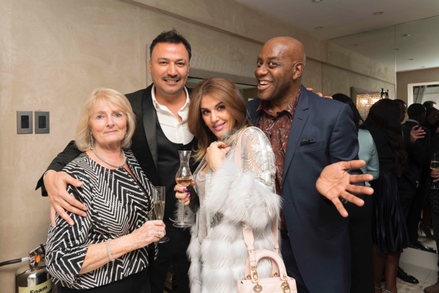 Four people posing together at a social event, dressed in formal attire. One woman is wearing a patterned dress, and another is in a light, fur-lined coat, holding a purse. Two men are smiling, one in a black suit and the other in a dark blazer, holding a glass.