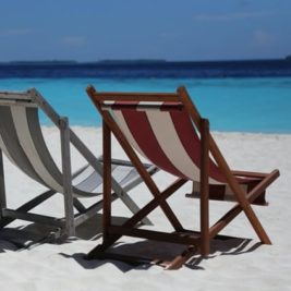 Two empty deck chairs, one grey and one red and white striped, sit on a pristine white sandy beach facing a calm, azure ocean under a clear blue sky. The distant horizon shows a thin line of low-lying islands or land masses.