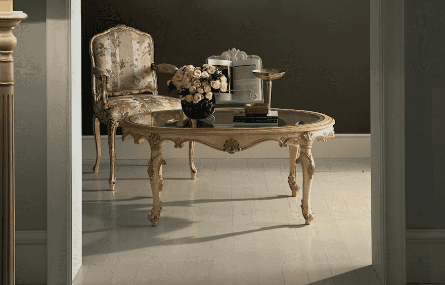 An elegant room with a vintage armchair, a glass-top ornate wooden table holding a black vase with white roses, and a silver decorative item. The room has a polished wooden floor, muted wall color, and classical furnishings creating a sophisticated ambiance.