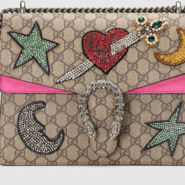 This stylish handbag with a beige and brown Gucci pattern, adorned with colorful, bedazzled patches including stars, moons, a heart pierced by a sword, and a crystal-studded horseshoe clasp, is perfect for luxury Valentine's gifts. The bag features a silver chain strap and a pink flap accent.