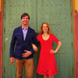 A man and woman stand next to each other in front of a large green, ornate door with peeling paint. The man wears a blue jacket and khaki pants, while the woman wears a red dress with a unique cutout pattern at the hem. Both are smiling. The background includes red walls.