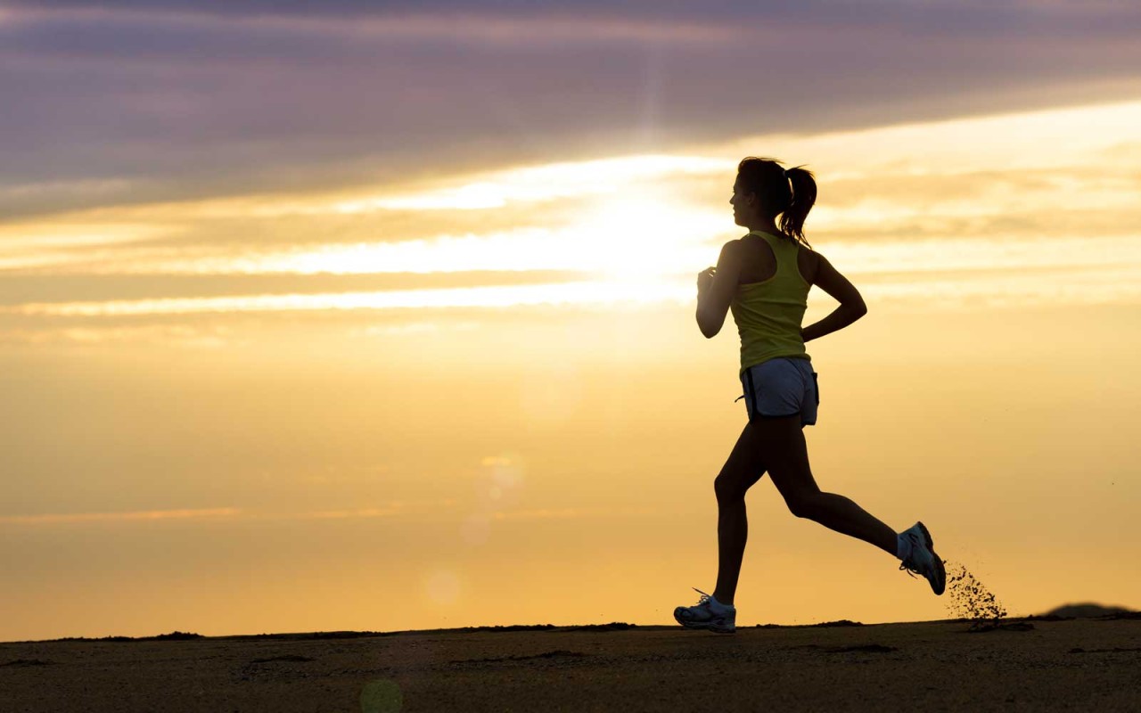 A person is jogging along a beach at sunset, with bright orange and yellow hues in the sky. The runner is in silhouette, wearing a tank top and shorts. The setting sun casts a warm glow, creating a serene atmosphere—perfect for winding down before learning how to get the best night’s sleep.