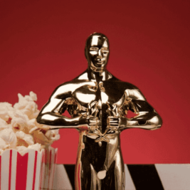 A gold statue resembling an Oscar award stands prominently against a red background. To the left of the statue is a container of popcorn with red and white stripes. The bottom portion of the image features a black and white checkered pattern.