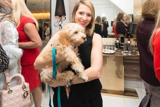 A woman in a black dress is smiling and holding a fluffy, light-brown puppy with a blue leash. She is standing in a well-lit room with other people in the background, some dressed in red and some talking. There is a gold-colored bar area with bottles and glasses behind her.