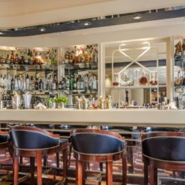 A modern American bar with a well-stocked selection of spirits arranged on shelves behind the counter. Four leather bar stools line the sleek bar top, which is adorned with various bottles, shakers, and barware. Mirrors and lights add a polished touch to the sophisticated setting.