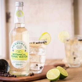 A bottle of Fentimans Botanical Tonic Water sits on a wooden surface next to a glass filled with ice cubes, tonic water, and a slice of lime, perfect for refreshing mocktails. Juniper berries are scattered on the surface, with a sliced lime in the foreground.