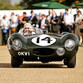 A vintage race car with the number 14 and a "OKVI" license plate drives down a gravel path in front of an audience. People are gathered on both sides, some taking photos, under green umbrellas and a clear sky. The driver is focused ahead while driving.