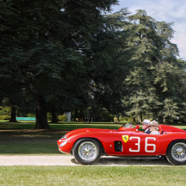 A red vintage racing car with the number 36 and the Ferrari logo on the side drives along a dirt path in front of a large mansion surrounded by trees on a sunny day. The driver and passenger are wearing helmets.