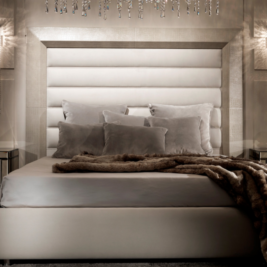 A luxurious bedroom, perfect for those *choosing a luxury bed*, featuring a large upholstered bed with multiple grey and silver pillows and a furry blanket. Elegant pendant lights hang on either side, while mirrored nightstands flanking the bed add a touch of sophistication to the room.