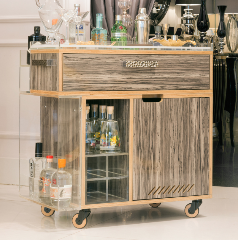 A stylish wooden bar cart on wheels features a top drawer with a handle, an array of bottles, glasses, and bar tools on the surface. The cart has compartments holding various liquor bottles and space for additional storage. It stands in a modern, elegant room.