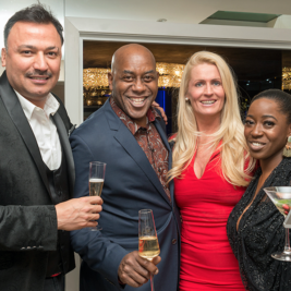 Four people are posing and smiling at a social event, each holding a drink. The man on the left is in a black suit, the man in the middle is in a patterned shirt, the woman next to him is in a red dress, and the woman on the right is holding a martini glass and a phone.