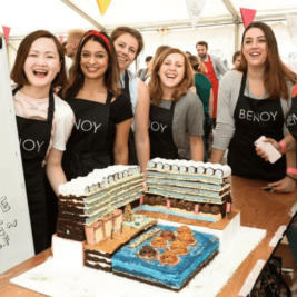 A group of six women, all wearing black aprons with the word "BENOY" on them, are smiling and posing behind a detailed architectural cake at an event. The cake resembles a modern building complex. A sign next to the cake reads "TASTE ME" with additional text underneath.