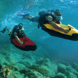 Two divers, each using an underwater scooter, explore a rocky, coral-rich seabed. They wear snorkeling masks and breathing apparatus. The scooter on the right is yellow, and the one on the left is red. The water is clear, providing a bright, vibrant view of the underwater scene.