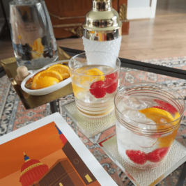 Two cocktails with ice, orange slices, and raspberries are on coasters atop a glass table, accompanied by a dish of orange slices, a bottle of tonic water, and a shaker. Nearby, a colorful illustration of a building is resting on the table. A patterned rug is visible below.