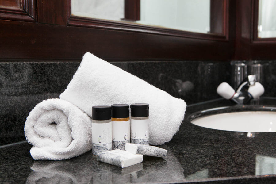 A neatly rolled white towel, three small bottles of toiletries, two wrapped items, and an unfolded towel are arranged on a dark granite bathroom counter. A sink with a countertop and a mirror with a wooden frame are visible in the background, ensuring you can be the perfect host.