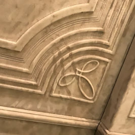 A close-up of an ornate ceiling corner features intricate raised designs and moldings, reminiscent of the opulent styles showcased at Salone del Mobile Milano. The patterns include intersecting curves and loops, highlighting textured details in a light beige hue.