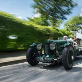 A vintage green sports car speeds down a suburban street lined with greenery and houses. The driver, wearing sunglasses, is focused on the road ahead. The car's classic design features exposed wheels and a sleek, aerodynamic body. Bright, sunny day with a clear blue sky.