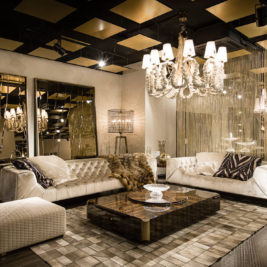 A luxurious living room features two plush cream sofas adorned with cushions and a fur throw. Two large mirrors, chandeliers, and warm lighting enhance the elegant ambiance. A glass coffee table sits atop a geometric patterned rug, and a quilted ottoman completes the setting. .