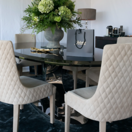 A glass dining table with a large floral centerpiece is surrounded by cream-colored, quilted chairs. On the table, there is a shopping bag with “Juliet” printed on it. In the background, a sideboard holds a coffee machine and various supplies.