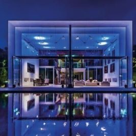 A modern glass-fronted house illuminated with blue and purple lights, reflecting off a tranquil swimming pool in the foreground. The interior is visible, showcasing contemporary furnishings. Trees and well-lit landscaping surround the property under a night sky.